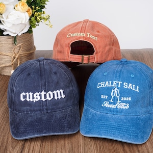 May include: Three baseball caps with different designs. One is a light orange cap with the text "Custom Text" embroidered on the front. Another is a dark blue cap with the word "custom" embroidered in white on the front. The third is a dark blue cap with the text "CHALET SALI EST 2025 Social Club" embroidered in white on the front.