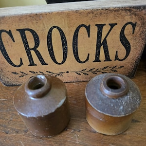 May include: Two small, cylindrical brown stoneware crocks with narrow openings at the top. Behind the crocks sits a rectangular wooden sign with a distressed tan finish and the word CROCKS printed in large, bold, black capital letters.