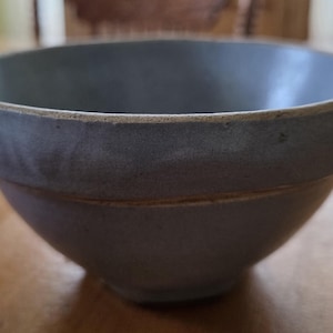 May include: A round ceramic bowl with a smooth, matte light blue glaze on the interior and exterior. The bowl features a thick, rounded rim with an exposed, unglazed tan clay edge that contrasts with the blue finish of the vessel body.