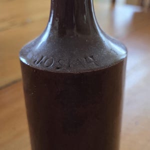 May include: A dark brown, cylindrical glass bottle with a narrow neck and a rounded rim. The shoulder of the bottle features embossed lettering that reads JOSIAH. The surface of the glass has a smooth, reflective finish with a deep, opaque colour.