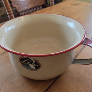 May include: A cream-coloured enamel basin with a red rim and a single side handle. The exterior features a dark label with the text London Enamel and Made in China. The interior is smooth and cream-coloured, matching the exterior finish of the bowl.