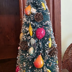May include: A conical bottle brush tree in dark green with a dusting of silver glitter. The tree is decorated with small brown pinecones, miniature red and yellow fruit ornaments, and silver leaves, all mounted on a round red base.