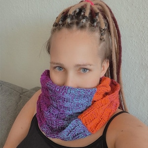 Könnte beinhalten: Ein gestrickter Loop-Schal in Lila, Blau und Orange. Der Schal ist aus dickem Garn mit einem strukturierten Muster gefertigt. Der Schal ist um den Hals gewickelt.