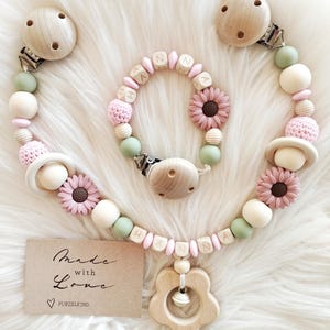Könnte beinhalten: Ein Set Baby-Accessoires, darunter ein Schnullerclip, ein Armband und eine Halskette, mit Holzperlen, rosa und grünen Akzenten und Blumendetails. Der Schnullerclip und die Halskette sind mit einem blütenförmigen Beißring aus Holz verbunden. Auf einer kleinen Karte steht "Made with Love".