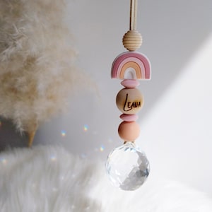 May include: A hanging rainbow-themed car charm with a crystal ball at the bottom. The charm features a wooden rainbow, wooden beads, and the name "LEAH" engraved on a wooden bead. The charm is suspended by a beige cord.