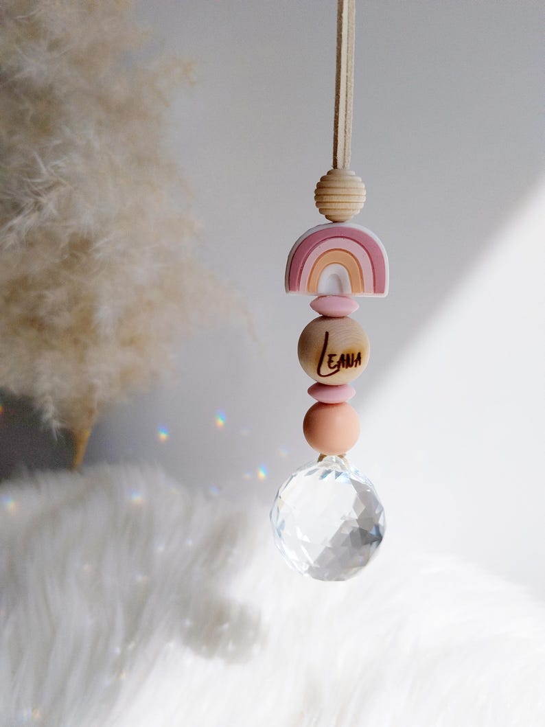 May include: A hanging rainbow-themed car charm with a crystal ball at the bottom. The charm features a wooden rainbow, wooden beads, and the name "LEAH" engraved on a wooden bead. The charm is suspended by a beige cord.