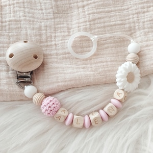 Könnte beinhalten: Ein Baby-Schnullerclip mit dem Namen "ALENA" aus Holzbuchstaben-Perlen. Der Clip hat einen Holzclip, weiße und rosa Perlen, eine rosa gehäkelte Perle und eine weiße blütenförmige Perle. Der Schnullerclip liegt auf einer weißen, flauschigen Oberfläche.