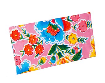 May include: A rectangular, pink fabric item with a vibrant floral pattern. The design features large blue, red, and yellow flowers with green leaves. The item is likely a wallet or small pouch, showcasing a colorful, botanical theme.