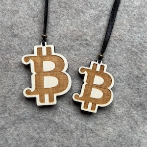 Könnte beinhalten: Zwei Holzanhänger mit Bitcoin-Symbol, hellbraunes Design und schwarzes Band. Die Anhänger sind aus Holz und haben eine natürliche Oberfläche. Das Bitcoin-Symbol ist ein stilisiertes "B" mit vertikalen Linien.
