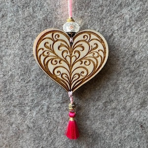 Könnte beinhalten: Holzherz-Ornament mit aufwendigem Schnitzmuster. Es hängt an einer rosa Schnur und hat eine transparente Perle, goldene Akzente und eine leuchtend pinkfarbene Quaste. Das Herz hat eine natürliche Holzfarbe.