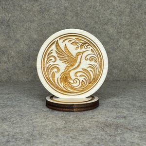 Könnte beinhalten: Ein runder Holzuntersetzer mit einem lasergeätzten Kolibri-Design. Der Kolibri ist von wirbelnden Linien und Blättern umgeben. Der Untersetzer steht auf einem kleineren, dunkleren Holzsockel. Der Untersetzer hat einen Durchmesser von etwa 10 cm.