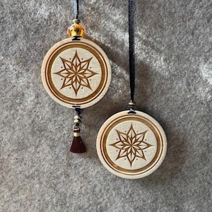 Könnte beinhalten: Zwei runde Holzornamente mit Sternmuster. Die hellbraunen Ornamente haben einen dunkelbraunen Rand und Stern. Eines hat eine braune Quaste und goldene Perlen. Beide hängen an schwarzen Bändern.