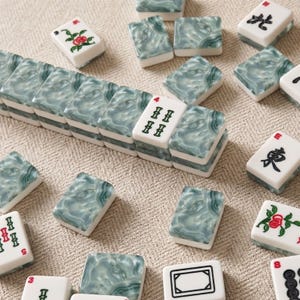 Puede incluir: Un conjunto de fichas de mahjong rectangulares con una base blanca y un patr&oacute;n jaspeado de remolinos verdes y grises en el reverso. Algunas fichas muestran caracteres chinos negros, motivos florales rojos o s&iacute;mbolos de bamb&uacute; verdes con n&uacute;meros rojos.