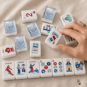 May include: A set of rectangular mahjong tiles featuring winter-themed illustrations including skiers, a lift ticket, mittens, a scarf, and snowflakes. Some tiles have a clear, icy blue textured surface, while others display colorful graphics and text.