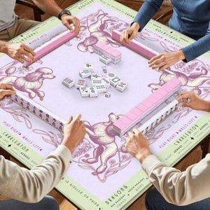 May include: A square Mahjong game table with a light purple surface featuring a poodle illustration. Pink tile racks and white tiles are arranged around the table. Players are seen interacting with the game pieces.