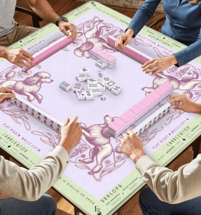 May include: A square Mahjong game table with a light purple surface featuring a poodle illustration. Pink tile racks and white tiles are arranged around the table. Players are seen interacting with the game pieces.