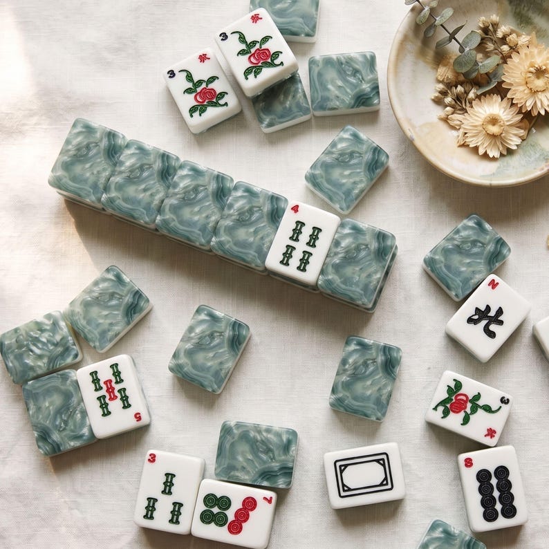 Puede incluir: Un conjunto de fichas de mahjong rectangulares con un reverso de m&aacute;rmol verde salvia y un frente blanco con varios s&iacute;mbolos grabados en negro, rojo y verde, incluyendo patrones florales, caracteres chinos y puntos num&eacute;ricos.