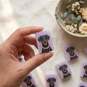May include: A rectangular tile featuring a printed illustration of a black and tan Doberman dog wearing a pink beaded collar. The tile has a smooth, light purple background and is held between fingers, with other identical tiles scattered nearby.