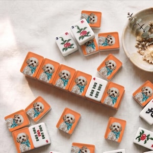 May include: A collection of rectangular mahjong tiles featuring custom printed images of a fluffy white dog wearing a blue bandana. Some tiles display traditional mahjong symbols in red, green, and black, while others show the dog portrait.