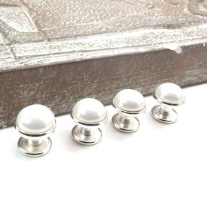 May include: Four silver and white pearl shirt studs. The studs are round and have a smooth, shiny finish.