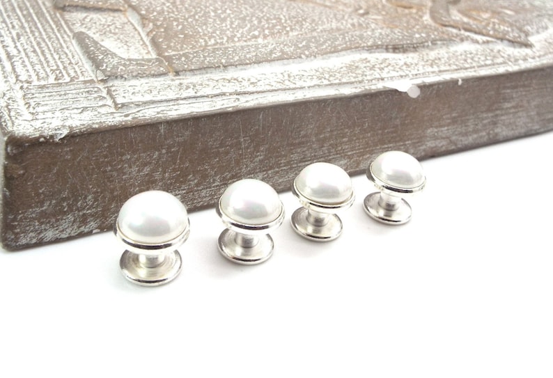 May include: Four silver and white pearl shirt studs. The studs are round and have a smooth, shiny finish.
