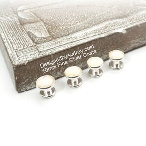 May include: Four silver dome-shaped buttons with a white pearl-like finish. The buttons are 10mm in diameter and are arranged in a row on a white surface. The text "DesignedbyAudrey.com 10mm Fine Silver Dome" is visible on a brown wooden surface in the background.