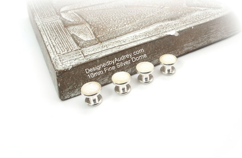 May include: Four silver dome-shaped buttons with a white pearl-like finish. The buttons are 10mm in diameter and are arranged in a row on a white surface. The text "DesignedbyAudrey.com 10mm Fine Silver Dome" is visible on a brown wooden surface in the background.