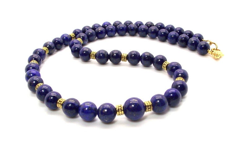 May include: A necklace featuring deep blue, spherical beads. The beads are separated by small, gold-colored spacers. The necklace is displayed against a white background, highlighting the contrast between the colors. The clasp is gold-colored.