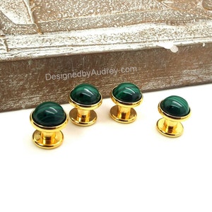 May include: Four gold-toned cabinet knobs with malachite green stone tops. The knobs are displayed on a white surface with a textured wooden background. The website DesignedbyAudrey.com is visible in the background.