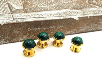 May include: Four gold-toned cabinet knobs with malachite green stone tops. The knobs are displayed on a white surface with a textured wooden background. The website DesignedbyAudrey.com is visible in the background.
