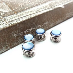 Puede incluir: Cuatro botones de camisa plateados con tapas de nácar azul y blanco. Los botones están dispuestos en semicírculo sobre una superficie blanca. El texto "DesignedbyAudrey.com" es visible en una superficie de madera en el fondo.