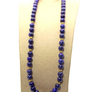 May include: A long necklace featuring large, round, deep blue beads. The beads are separated by gold-colored spacers. The necklace is displayed on a neutral-toned mannequin, showcasing its length and design.