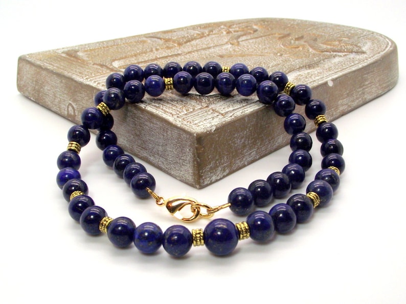 May include: A necklace featuring deep blue, spherical beads. The beads are strung together with gold-colored accents and a gold clasp. The necklace is displayed on a light-colored, textured surface.