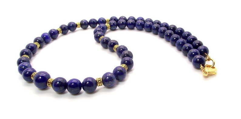 May include: A necklace featuring deep blue, spherical beads. The beads are separated by gold-colored spacers and are strung together. The necklace is secured with a gold-colored clasp. The jewelry is displayed against a white background.
