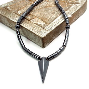 May include: A black hematite necklace with a pointed hematite pendant. The necklace is made of round beads and is lying on a wooden surface.