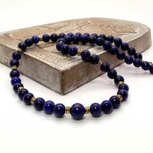 May include: A dark blue beaded necklace with gold-colored accents. The necklace features round, polished beads and small gold-colored spacers. The necklace is displayed on a light brown, carved wooden surface against a white background.