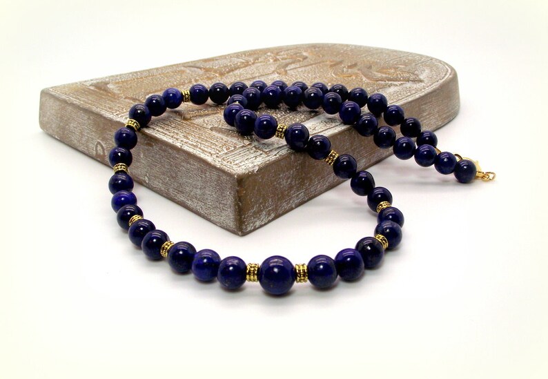 May include: A dark blue beaded necklace with gold-colored accents. The necklace features round, polished beads and small gold-colored spacers. The necklace is displayed on a light brown, carved wooden surface against a white background.