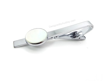 May include: Silver tie clip with a round, white, iridescent button. DesignedbyAudrey.com is printed on the clip.