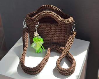 May include: A brown crocheted handbag with a matching shoulder strap and silver-tone hardware. The bag has a small handle and a decorative charm with a flower and a green tag. The bag is sitting on a white surface.