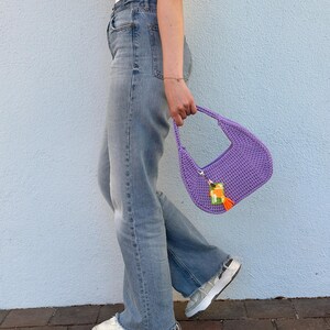 May include: A purple crocheted handbag with a tassel and a colorful charm. The bag is being held by a person wearing blue jeans and white sneakers.