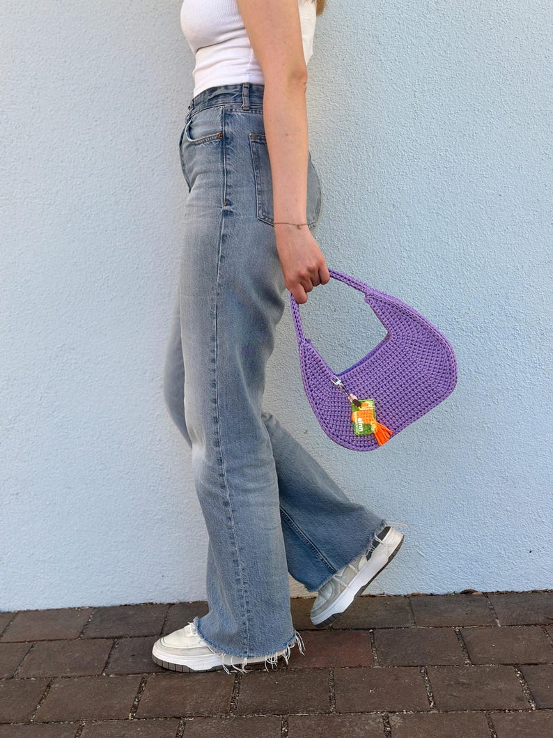 May include: A purple crocheted handbag with a tassel and a colorful charm. The bag is being held by a person wearing blue jeans and white sneakers.