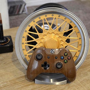 May include: A brown Xbox controller with a wood grain finish rests on a silver stand. Behind it is a gold and silver car wheel rim. The controller has black buttons and a black directional pad. The rim has a gold center and gold accents.