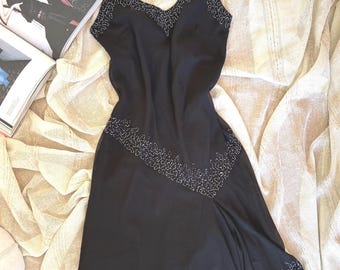 May include: A black slip dress with delicate spaghetti straps and a V-neckline. The dress is adorned with small, silver-colored beads along the neckline and hem. The dress is made of a flowing, lightweight fabric.
