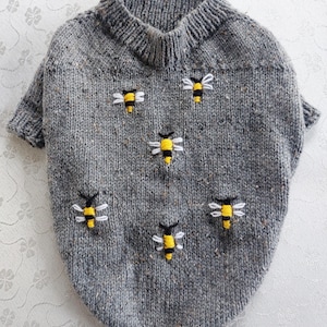 May include: A grey speckled knit sweater featuring a ribbed collar and short sleeves. The sweater is decorated with five embroidered bees, each designed with yellow and black striped bodies and white thread wings, scattered across the front.
