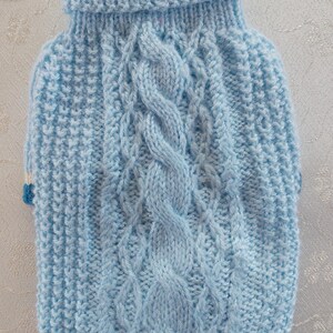 May include: A light blue knitted jumper featuring a central vertical cable knit pattern flanked by textured stitch panels. The garment has a ribbed turtleneck collar with horizontal white and dark blue stripes at the top edge.