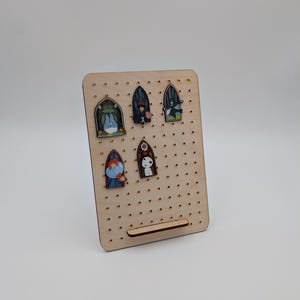 May include: A wooden pin display board with multiple pins attached. The pins feature colorful illustrations of characters in a stained-glass window design. The board has a rectangular shape with a small shelf at the bottom.