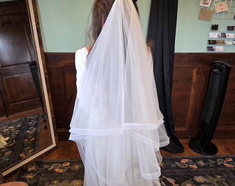 May include: A white wedding dress with a layered veil. The veil is made of a sheer fabric and falls to the shoulders. The dress is long and flowing, with a simple design. The background includes a mirror, a rug, and a black curtain.
