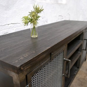 May include: A dark-stained wooden media console with metal mesh door panels and handles. The top surface is smooth and dark, with a small glass vase holding green flowers. The cabinet has open shelving and a modern industrial design.