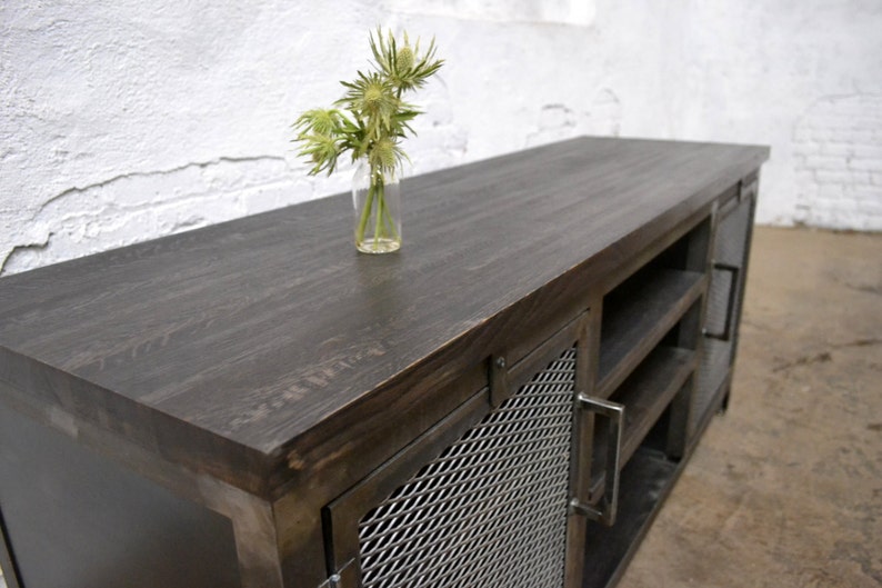 May include: A dark-stained wooden media console with metal mesh door panels and handles. The top surface is smooth and dark, with a small glass vase holding green flowers. The cabinet has open shelving and a modern industrial design.