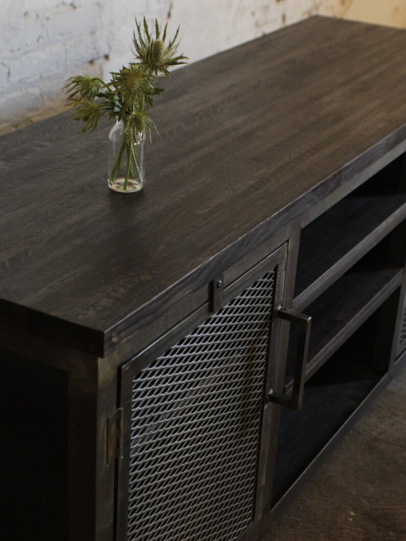 May include: A dark-stained wooden media console with a metal mesh door and open shelving. A small glass vase with green thistle-like flowers sits on top. The console has a modern, industrial design.